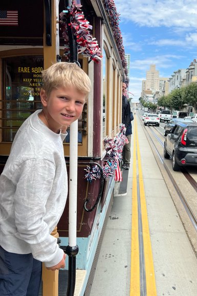 Ein Junge mit blonden Haaren lächelt, während er an einem historischen Straßenbahnwagen in San Francisco steht.