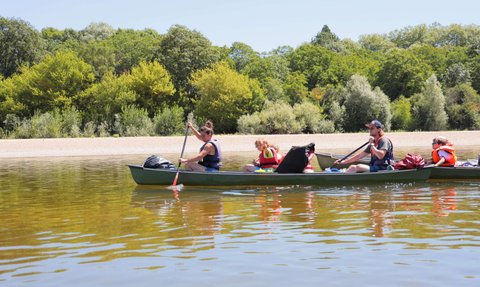 Eine Gruppe von vier Personen paddelt in einem Kanu auf einem ruhigen Fluss, umgeben von üppigem Grün und klarem Himmel.