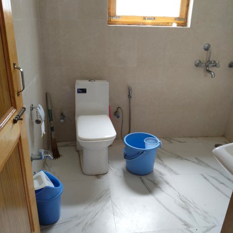 Ein einfaches Badezimmer mit einer Toilette, zwei blauen Eimern und einem Fenster, das natürliches Licht hereinlässt.