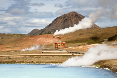 Ein geothermisches Kraftwerk mit rauchenden Schornsteinen vor einer beeindruckenden Berglandschaft in Island.