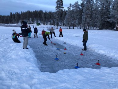 Eine Gruppe von Menschen spielt Eisstockschießen auf einer schneebedeckten Fläche, umgeben von hohen, frostigen Bäumen.
