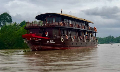 Ein traditionelles Holzboot mit rotem Bug schwebt sanft auf dem ruhigen Wasser, umgeben von üppigem Grün und bewölktem Himmel.