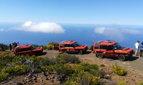 Drei orangefarbene Geländewagen stehen auf einem schmalen Weg mit Blick auf das blaue Meer und die Wolken darüber.