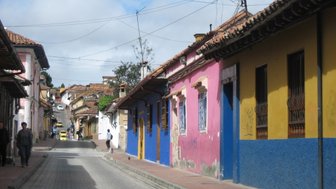 Bunte Häuser säumen die Straße in Bogotá, während Passanten und ein gelbes Taxi vorbeifahren.