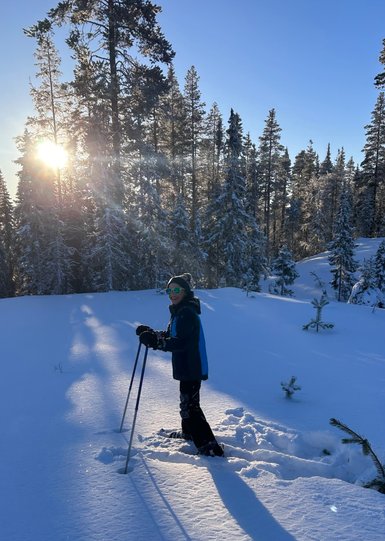Ein fröhliches Kind steht im tiefen Schnee, umgeben von schneebedeckten Bäumen, während die Sonne im Hintergrund scheint.