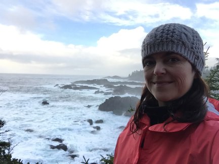 Eine Frau mit einem grauen Hut lächelt vor einer stürmischen Küstenlandschaft in Tofino, umgeben von Wellen und Felsen.