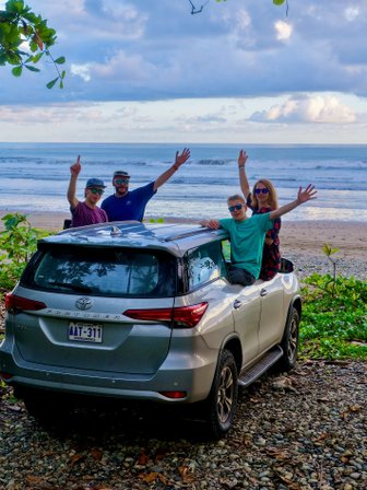 Eine Gruppe von vier Personen steht fröhlich neben einem silbernen Auto am Strand von Costa Rica, umgeben von üppigem Grün.