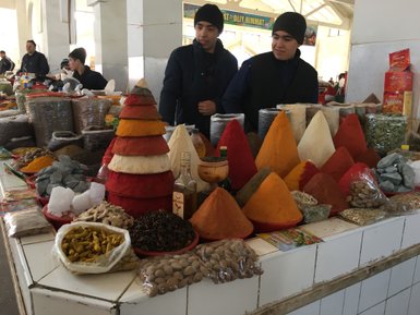 Zwei Männer stehen an einem Marktstand, umgeben von bunten Gewürzen in verschiedenen Formen und Farben.