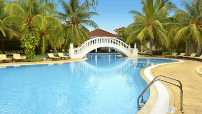 Ein malerischer Pool mit einer eleganten weißen Brücke, umgeben von üppigen Palmen und Sonnenliegen, strahlt tropisches Flair aus.