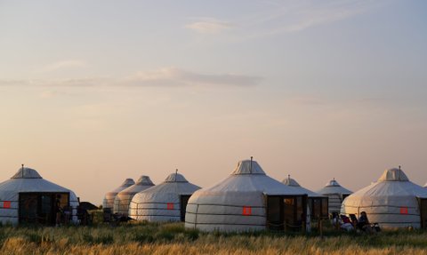 In der sanften Abenddämmerung stehen mehrere Jurten in einer weiten Graslandschaft, umgeben von sanften Hügeln.