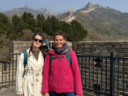 Zwei Frauen stehen lächelnd auf der Chinesischen Mauer, umgeben von grünen Bäumen und einer beeindruckenden Berglandschaft.