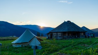Ein Zelt steht auf einer Wiese, während die Sonne hinter den Bergen aufgeht und ein Holzhaus im Hintergrund sichtbar ist.