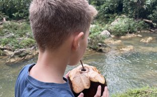 Ein Junge steht am Ufer eines klaren Flusses und hält eine frische Kokosnuss in der Hand, umgeben von üppigem Grün.