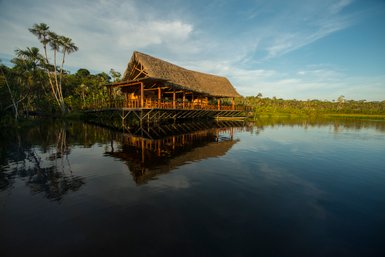 Ein malerisches Holzhaus mit Strohdach steht am Ufer eines ruhigen Gewässers, umgeben von üppiger tropischer Vegetation.