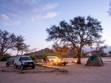 Ein Campingplatz in Namibia mit Zelten, einem Lagerfeuer und Fahrzeugen unter einem großen Baum in der Dämmerung.