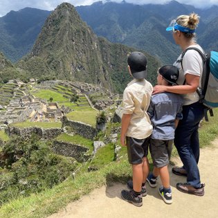 Eine Frau steht mit zwei Kindern an einem Aussichtspunkt und bewundert die Ruinen von Machu Picchu in der Ferne.