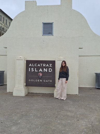Eine Person steht vor dem Alcatraz-Schild, umgeben von einer grauen, bewölkten Atmosphäre und historischen Gebäuden im Hintergrund.