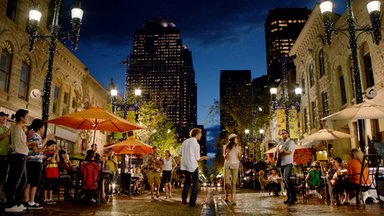 Eine belebte Straße in Calgary bei Nacht, mit Menschen, die an Tischen sitzen und sich unterhalten, umgeben von Lichtern.
