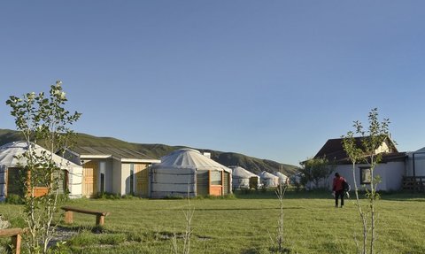 Eine Gruppe von Jurten steht in einer grünen Wiese, umgeben von sanften Hügeln unter einem klaren blauen Himmel.