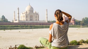 Eine Frau sitzt am Ufer und betrachtet das majestätische Taj Mahal, umgeben von einer ruhigen Landschaft.