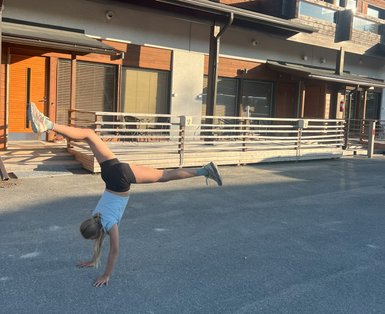 Ein Mädchen macht einen Handstand auf einer Straße vor modernen Holzhäusern, während die Sonne scheint.