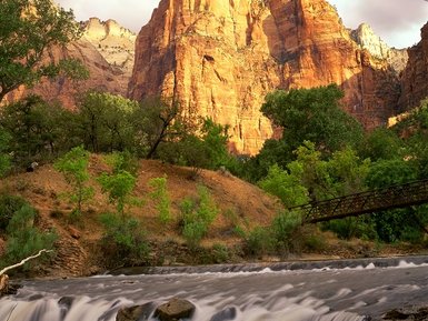 Ein majestätischer, rotbrauner Felsen erhebt sich über einen ruhigen Fluss, umgeben von üppigem Grün und sanften Wolken.