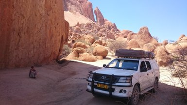 Ein Junge sitzt neben einem Jeep mit Dachzelt im Schatten eines Felsens - Namibia Familienreise