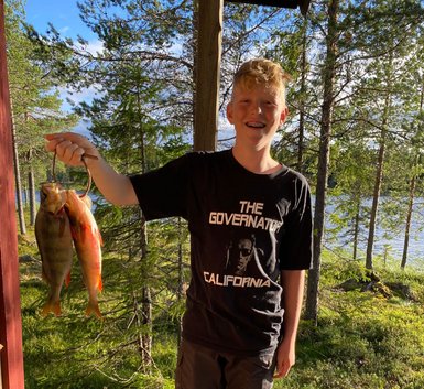 Ein Junge steht auf einer Holzveranda und hält stolz zwei gefangene Fische in der Hand, umgeben von Bäumen und Wasser.