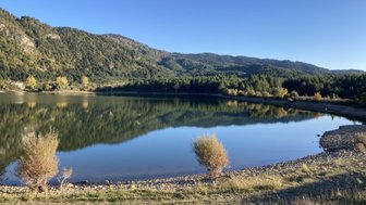 Ein ruhiger See spiegelt die umliegenden Berge und Bäume wider, während die Sonne am klaren Himmel scheint.