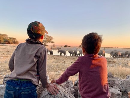 Zwei Kinder beobachten fasziniert eine Gruppe von Elefanten, die am Wasserloch in Namibia trinken, während die Sonne untergeht.