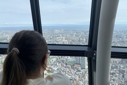Eine Person mit einem Pferdeschwanz blickt aus dem Fenster des Tokio Skytree auf die weitläufige Stadtlandschaft.