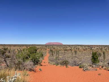 Im Vordergrund erstreckt sich ein Weg aus rotem Sand, während im Hintergrund der markante Uluru-Einschnitt zu sehen ist.