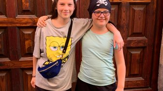 Zwei Mädchen stehen lächelnd vor einer Holztür, eines trägt ein Spongebob-T-Shirt und das andere ein Piratenkopftuch.