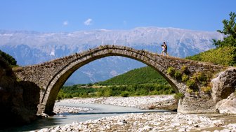 Ein Paar geht über eine alte Steinbrücke, umgeben von grünen Hügeln und majestätischen Bergen unter einem strahlend blauen Himmel.