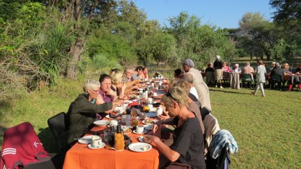 Eine Gruppe von Menschen genießt ein gemeinsames Frühstück im Freien, umgeben von üppiger Natur und Sonnenschein.