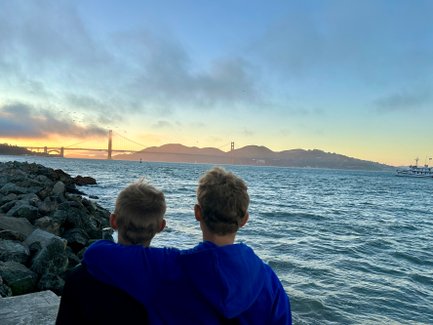 Zwei Jungen stehen am Wasser und blicken auf die Golden Gate Bridge, während die Sonne hinter den Hügeln untergeht.