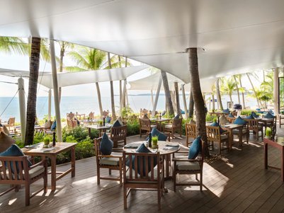 Ein elegantes Restaurant am Strand mit Holztischen und Stühlen, umgeben von Palmen und Blick auf das glitzernde Wasser.