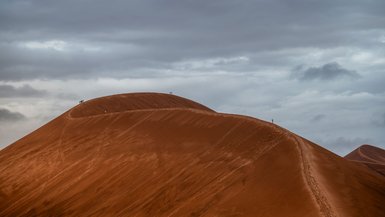 Wüstenlandschaft mit roten Sanddünen unter einem bewölkten Himmel, Wanderer erklimmen die hohe Sandstelle.