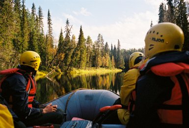 Eine Gruppe von Menschen in einem Schlauchboot, umgeben von hohen Bäumen und einem ruhigen Fluss in Finnland.