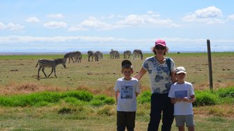 Eine Frau steht mit zwei Kindern auf einer Wiese, während Zebras im Hintergrund grasen und der Himmel blau ist.