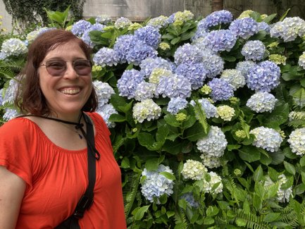 Eine lächelnde Frau mit braunen Haaren steht vor einem üppigen Hintergrund aus blühenden Hortensien in verschiedenen Farben.