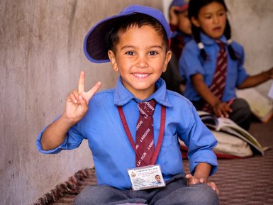 Ein fröhlicher Junge in Schuluniform sitzt auf dem Boden und zeigt ein Peace-Zeichen, während er ein Buch auf seinem Schoß hat.
