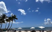 Ein sonniger Strand mit Palmen, Liegen und Sonnenschirmen, unter einem strahlend blauen Himmel mit wenigen Wolken.