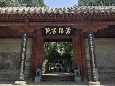 Ein traditionelles Tor mit roten Holzelementen und goldenen Schriftzeichen, umgeben von üppigem Grün und alten Steinen.