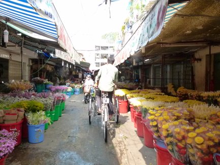 Ein belebter Markt in Saigon, gesäumt von bunten Blumenständen und Radfahrern, die durch die engen Gassen fahren.
