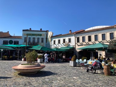 Ein belebter Platz mit Cafés und Restaurants, umgeben von historischen Gebäuden und Sonnenschirmen, unter strahlend blauem Himmel.