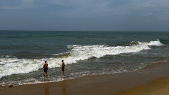 Zwei Personen stehen am Strand und beobachten die Wellen, während der Himmel leicht bewölkt ist und das Wasser glitzert.
