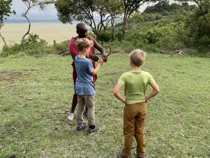 Ein Maasai zeigt zwei Jungen, wie man mit einem Bogen umgeht, während die grüne Landschaft im Hintergrund sichtbar ist.