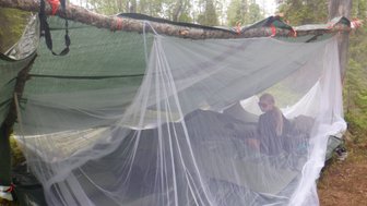 Eine Person sitzt in einem provisorischen Zelt, umgeben von einem Moskitonetz, in einer bewaldeten Umgebung.