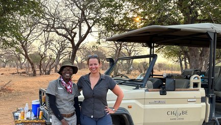 Zwei Personen stehen lächelnd vor einem Geländewagen in der afrikanischen Savanne, umgeben von kahlen Bäumen und sanftem Licht.